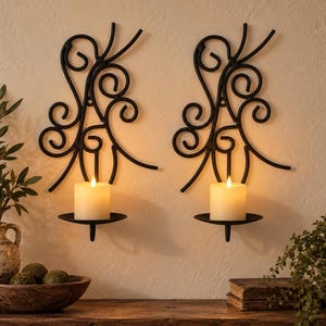 May include: A pair of black metal wall sconces featuring intricate scrollwork designs. Each sconce includes a circular platform holding a single white pillar candle. The metal frames are mounted vertically, displaying symmetrical curves and loops.