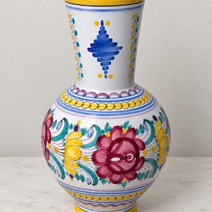 May include: A white ceramic vase with a rounded body and a flared rim. The surface is decorated with hand-painted floral motifs in magenta, yellow, and teal, accented by blue geometric patterns and horizontal bands of yellow and purple lines.