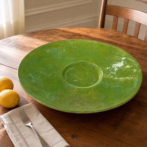 May include: A large, round, bright green ceramic serving platter with a glossy, mottled finish and a recessed circular center. The platter features subtle variations in color, ranging from lime to deep forest green, creating a textured appearance.