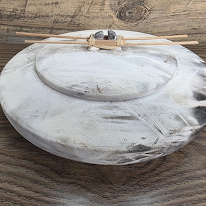 May include: A white and grey decorative bowl with a lid. The bowl has a distressed finish, with dark grey accents. Two wooden sticks are attached to the lid with a natural fibre. The bowl is on a wooden surface.