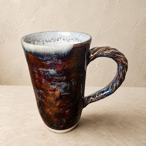 May include: A ceramic mug with a deep brown and black mottled glaze, featuring a white interior rim with a dripping effect. The mug has a distinctively textured, twisted rope-style handle in shades of blue and brown, creating a unique visual contrast.
