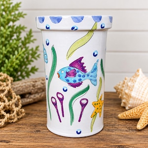 May include: A white cylindrical ceramic planter featuring a hand-painted underwater scene with a blue fish, a yellow starfish, green seaweed, purple coral, and blue bubbles. The rim is decorated with a repeating pattern of blue semi-circles.