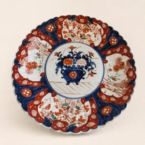 May include: A round ceramic plate with a scalloped edge, featuring a central blue and orange floral arrangement in a pot. The border is decorated with alternating panels of dark blue and orange floral patterns on a white background.