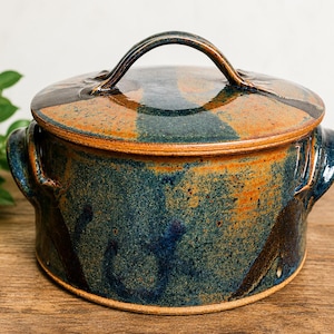 May include: A round ceramic casserole dish with a lid, featuring a mottled glaze in shades of deep blue, teal, and earthy orange. The pot has two side handles and a curved handle on the lid, with a smooth, glossy finish throughout the exterior.