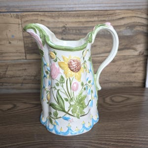 May include: A white ceramic pitcher with a floral design featuring sunflowers, pink and yellow flowers, and green leaves. The pitcher has a curved handle and a scalloped blue trim at the base. The rim is trimmed in green and pink.