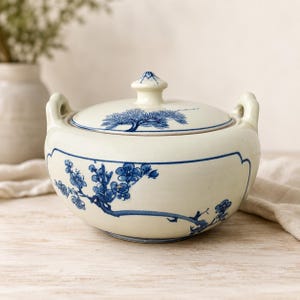 May include: A cream-colored ceramic lidded bowl with two side handles, featuring blue hand-painted floral branches on the body and a pine tree motif on the lid. The vessel has a smooth, glossy finish and a rounded shape with a decorative blue border.