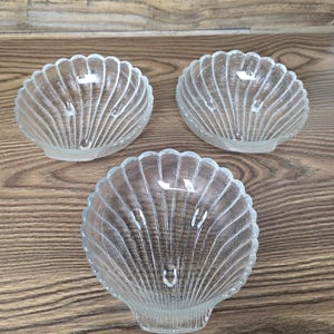 May include: Three clear glass seashell-shaped bowls. Each bowl has a scalloped edge and vertical ridged detailing. The bowls are arranged on a wooden surface.