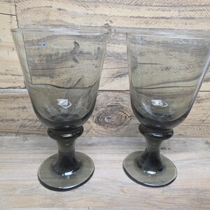 May include: Two gray-tinted stemmed glasses with a rounded base and a flared bowl. The glasses are made of a transparent material, allowing light to pass through. The stem has a bulbous design, and the base is oval-shaped.