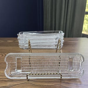 May include: Clear glass rectangular serving dishes with a textured surface. One dish is displayed on a gold-colored metal stand. A stack of similar dishes is visible in the background. The dishes are on a wooden table.