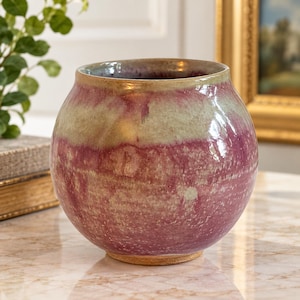 May include: A round ceramic vase featuring a glossy glaze with blended shades of magenta, deep pink, and muted cream. The vessel has a wide, curved body and a smooth, open rim, showcasing a reactive finish that creates a unique color gradient.
