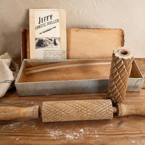 May include: A vintage Jiffy Cookie Roller set featuring a wooden rolling pin with a diamond-patterned texture, a separate cylindrical wooden roller with a matching diamond design, and a cardboard box with an instruction manual printed on the lid.