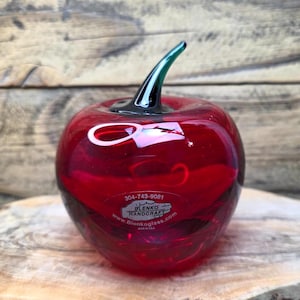 Może przedstawiać: Ręcznie robione, czerwone szklane jabłko z zieloną łodygą. Jabłko ma błyszczące wykończenie i stoi na drewnianej powierzchni. Jabłko ma naklejkę z napisem "BLENKO HANDCRAFT" i numerem telefonu.