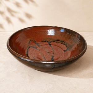 May include: A round ceramic bowl with a glossy finish, featuring a deep reddish-brown interior and a dark brown exterior. The interior surface displays a distinct, dark, abstract brushstroke pattern that contrasts with the warm clay tones.