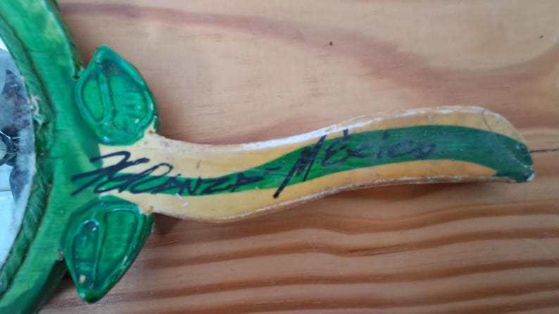Op de afbeelding: Een houten decoratief stuk met groene bladeren en een gele en groene streep. Het stuk heeft de handgeschreven tekst "Kalanza Mexico" in zwarte inkt. De achtergrond is een lichtbruin houten oppervlak.