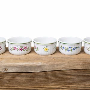 Puede incluir: Cinco ramequines de cerámica blanca con diseños florales y bordes verdes, colocados sobre una tabla de madera. Los diseños florales incluyen flores y bayas azules, rosas y amarillas. Cada ramequín mide unos 10 cm de diámetro.