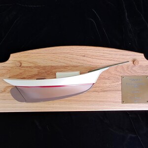 May include: A wooden sailboat model mounted on a light oak plaque. The boat is white with a red stripe and brown hull. A brass plaque with text is affixed to the right side. The model is a decorative nautical item.