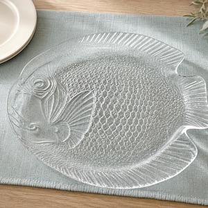 May include: A clear glass serving platter shaped like a fish, featuring a textured scale pattern on the body, defined fins, and a circular eye detail. The transparent plate has a smooth, glossy finish and a distinct outline resembling a marine fish.