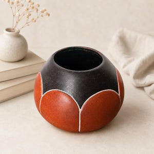 May include: A spherical ceramic vessel featuring a matte black upper section and a textured terracotta orange lower section. The design includes a repeating petal-like pattern defined by thin white lines that separate the black and orange areas.