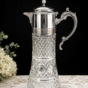 May include: A clear glass carafe with a diamond-cut pattern and a silver-toned metal top. The carafe features a tall, tapered body, a hinged lid with a small finial, a pointed spout, and an ornate, curved handle with decorative scrollwork.