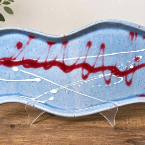 May include: A rectangular ceramic tray with wavy edges, featuring a light blue speckled glaze. The surface is decorated with a horizontal, wavy dark red painted line and thin, intersecting white paint splatters across the center.