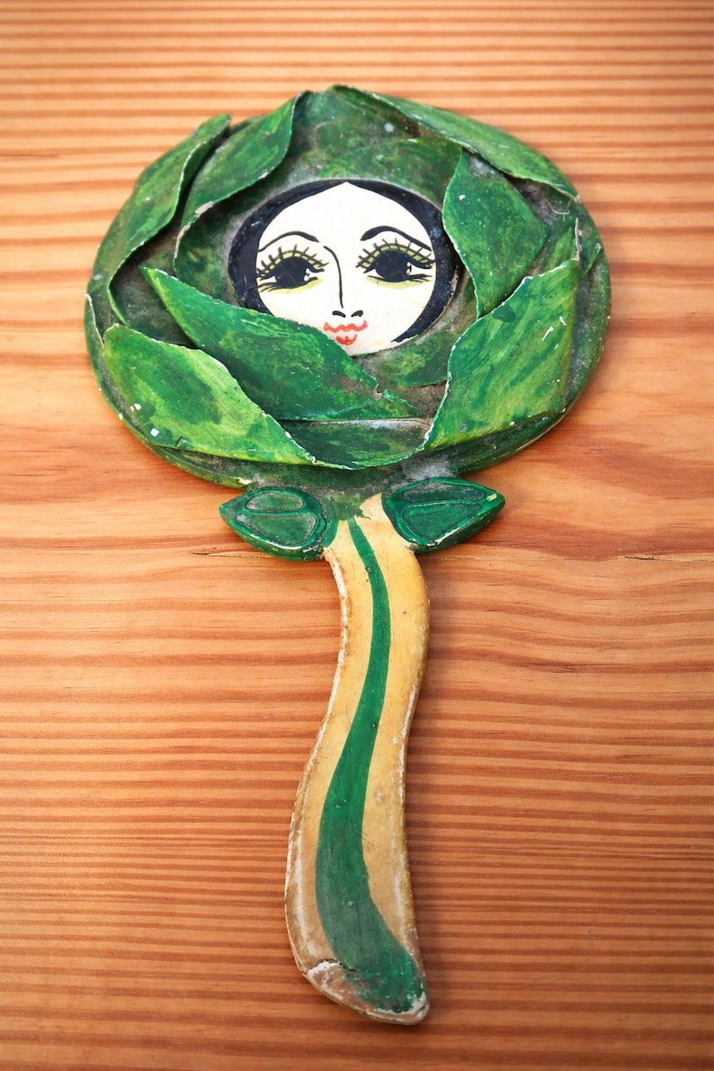 May include: A whimsical, hand-painted wooden wall hanging. The artwork features a face within a green leafy design, with a stem-like handle. The face has black hair, large eyelashes, and red lips. The stem is yellow with a green stripe.