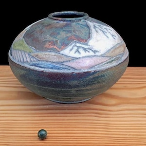 May include: A round ceramic vase with a landscape design featuring mountains and fields. The vase is primarily blue, green, and brown, with white accents on the mountains. A small, dark green marble sits on the wooden surface.