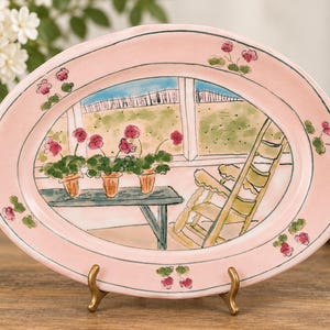 Puede incluir: Una bandeja ovalada de cerámica con un borde rosa claro y flores pintadas a mano. El centro muestra tres macetas con flores rosas sobre un banco azul junto a una mecedora amarilla, con vistas a un campo verde a través de una ventana.