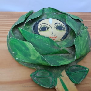 Op de afbeelding: Handgeschilderd houten decoratief stuk met een gezicht omringd door groene bladeren. Het gezicht heeft zwarte ogen, lange wimpers en een rode mond. De bladeren zijn in verschillende groentinten. Het stuk ligt op een houten oppervlak.