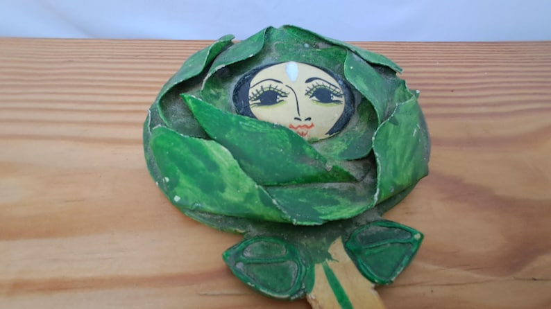 Op de afbeelding: Handgeschilderd houten decoratief stuk met een gezicht omringd door groene bladeren. Het gezicht heeft zwarte ogen, lange wimpers en een rode mond. De bladeren zijn in verschillende groentinten. Het stuk ligt op een houten oppervlak.