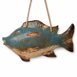 May include: A ceramic fish-shaped wall hanging with a blue and brown glaze. The fish has a textured surface with brown accents and a natural rope for hanging. The fish is approximately 12 inches long.