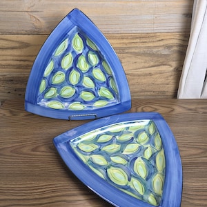 May include: Two blue, triangular serving platters with a hand-painted design. The interior of each platter features a pattern of green and yellow leaf-like shapes. The platters have a dark blue border and are displayed on a wooden surface.