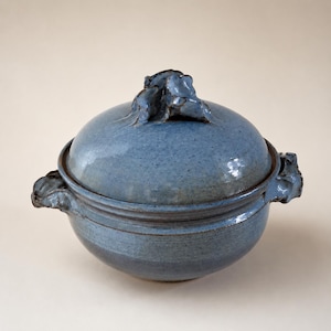 May include: A round ceramic lidded bowl with a speckled blue glaze. The lid features a sculptural, ruffled handle, and the bowl has two matching ruffled side handles. The surface has a glossy finish with subtle variations in the blue color tones.