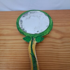Op de afbeelding: Handspiegel met een rond, reflecterend oppervlak. De lijst en het handvat van de spiegel zijn groen, geel en wit geschilderd, met bladachtige details aan de basis van de spiegelkop. De handgreep heeft een handtekening met zwarte inkt.