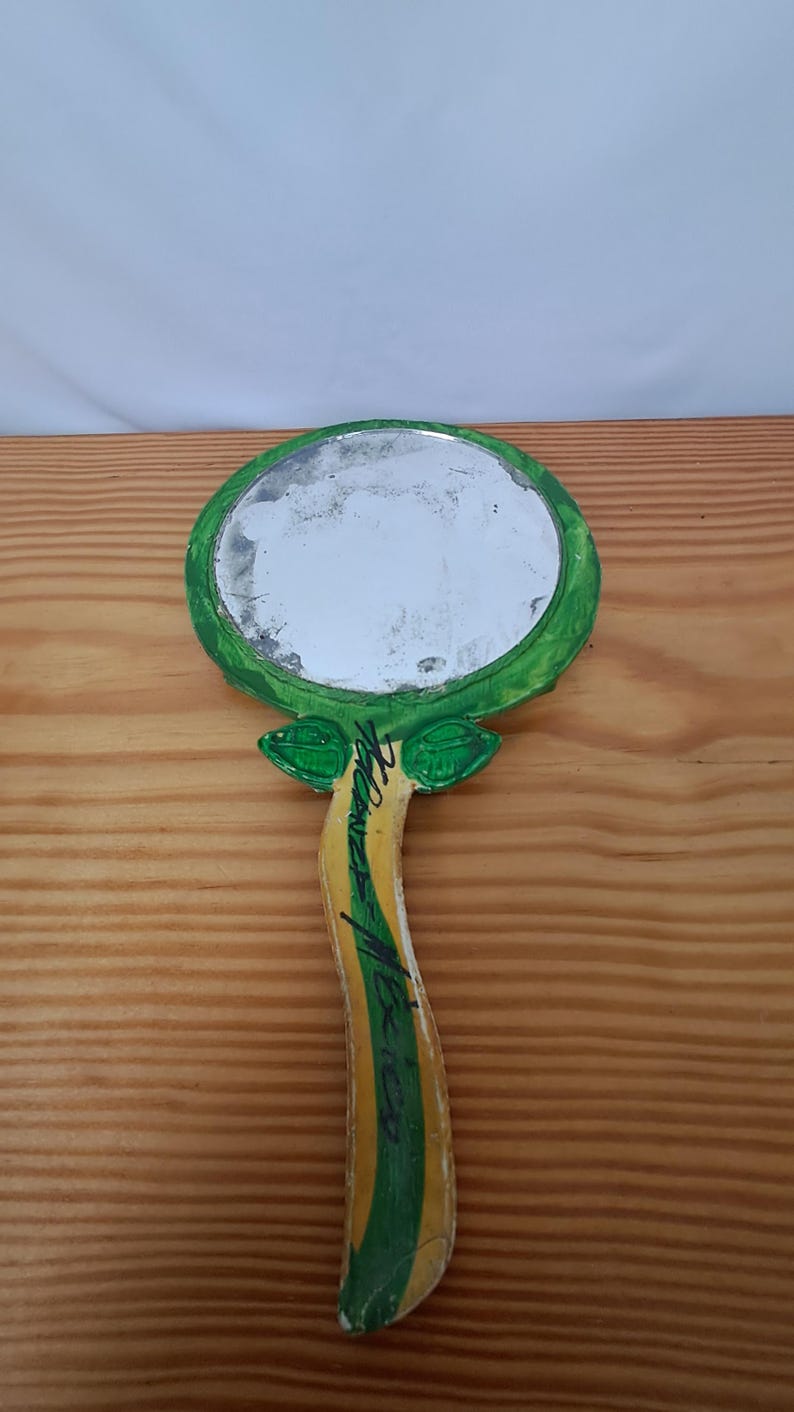 Op de afbeelding: Handspiegel met een rond, reflecterend oppervlak. De lijst en het handvat van de spiegel zijn groen, geel en wit geschilderd, met bladachtige details aan de basis van de spiegelkop. De handgreep heeft een handtekening met zwarte inkt.