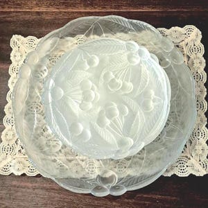 May include: Clear glass serving dishes with a cherry design. The set includes a large, scalloped-edge plate, a smaller plate, and a round dish. The dishes are placed on a white lace doily, with a dark wood background.