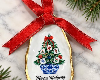 Op de afbeelding: Een oesterschelp ornament met een goudkleurige rand en een rood lint. De schelp is beschilderd met een kerstboom in een blauw-witte pot, versierd met Mahjong-tegels, kerstballen en de tekst Merry Mahjong.