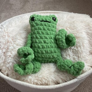 May include: A green crochet frog plush toy with a textured, chunky yarn finish. The frog features two large, round black eyes and a simple horizontal line for a mouth. It has long, flexible arms and legs with curled ends, sitting in a neutral position.