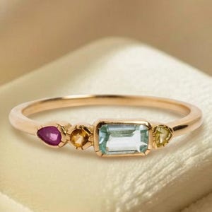 May include: A gold ring featuring a central rectangular light blue gemstone flanked by two small yellow stones and a single pear-shaped pink stone on one side. The band is thin and polished, showcasing a multi-stone arrangement in a horizontal line.