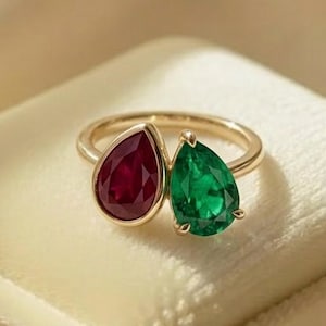 May include: A gold ring featuring two pear-shaped gemstones: a deep red ruby and a vibrant green emerald. The ring is set on a white velvet surface, with a soft, neutral background. The gemstones are the focal point, showcasing their color and cut.