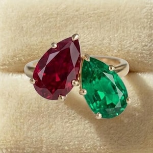 May include: A bypass ring featuring two pear-cut gemstones, one deep red ruby and one vibrant green emerald, set in a polished metal band. The stones are positioned side-by-side, showcasing their distinct teardrop shapes and faceted surfaces.
