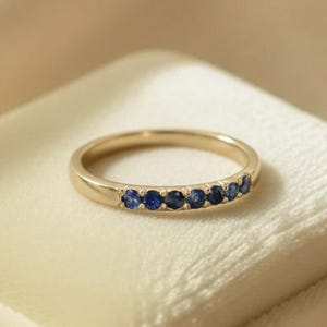 Puede incluir: Un anillo de banda dorada con una hilera de siete pequeñas piedras preciosas de zafiro azul oscuro, redondas, engastadas a lo largo de la mitad superior de la banda. El anillo se muestra sobre un cojín cuadrado de terciopelo color crema.