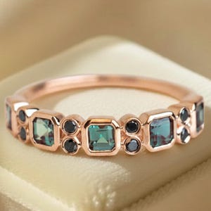 May include: Rose gold ring featuring alternating square and round gemstones. The square stones are teal, and the round stones are black. The ring is set on a cream-colored surface.