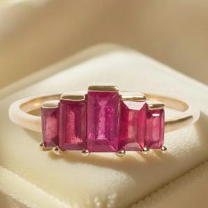 Può includere: Un anello d'oro con cinque pietre preziose rosa rettangolari. L'anello è presentato su un portagioie di velluto bianco, con uno sfondo morbido e neutro. Le pietre sono disposte a gradazione.