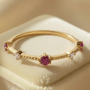 May include: A gold ring with a delicate design. The ring features a central ruby gemstone, flanked by smaller rubies and clear gemstones. The ring is set on a white velvet surface.