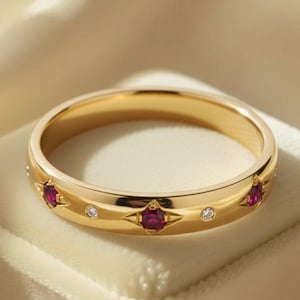 May include: A gold band ring with a series of alternating ruby and diamond accents. The ring is set on a white velvet surface, with a soft, neutral background.