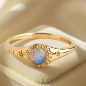 May include: A gold ring with a round, light blue and white stone at the center, surrounded by small clear stones. The band features star and crescent moon designs. The ring is set on a white velvet surface.