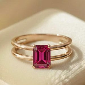 May include: A gold ring featuring a vibrant, emerald-cut pinkish-red gemstone set in the center. The band is designed with a double-wire structure that splits to hold the stone, creating a clean, polished look with a warm metallic finish.