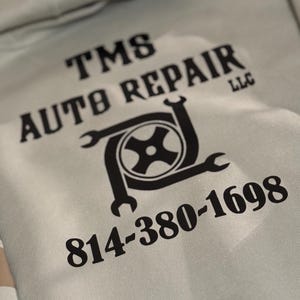 K&ouml;nnte beinhalten: Hellgrauer Kapuzenpullover mit schwarzem Text und Logo. Der Text lautet "TMS AUTO REPAIR LLC" und eine Telefonnummer. Das Logo zeigt einen Reifen und Schraubenschl&uuml;ssel.