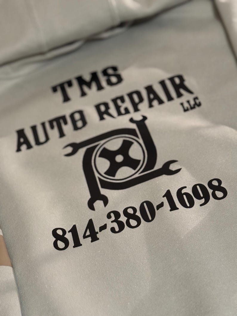 K&ouml;nnte beinhalten: Hellgrauer Kapuzenpullover mit schwarzem Text und Logo. Der Text lautet "TMS AUTO REPAIR LLC" und eine Telefonnummer. Das Logo zeigt einen Reifen und Schraubenschl&uuml;ssel.