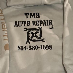 K&ouml;nnte beinhalten: Hellgrauer Kapuzenpullover mit dem Text "TMS AUTO REPAIR LLC" in schwarzen Blockbuchstaben. Darunter befindet sich eine Grafik eines Reifens und Schraubenschl&uuml;sseln. Die Telefonnummer 814-380-1698 ist darunter aufgedruckt. Das Carhartt-Logo ist auf dem &Auml;rmel zu sehen.